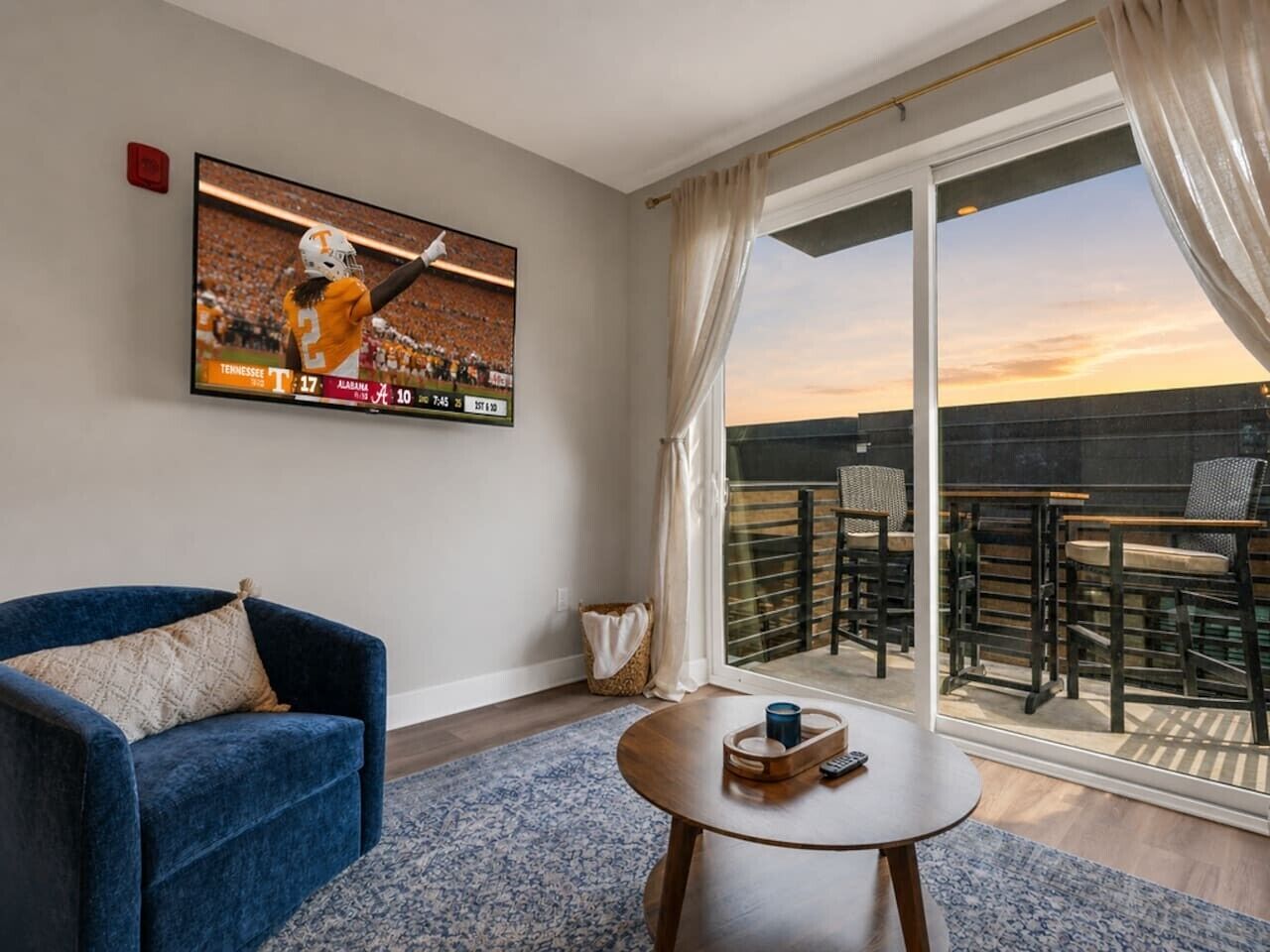 A room with a TV displaying a sports game, a blue armchair, a small round table with a tray, and a balcony with outdoor seating.