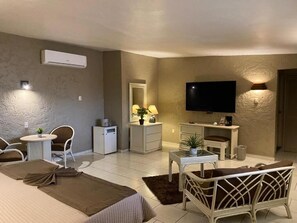 Habitación de hotel con cama, televisor, escritorio, silla, mesita, lámpara y equipo de aire acondicionado.