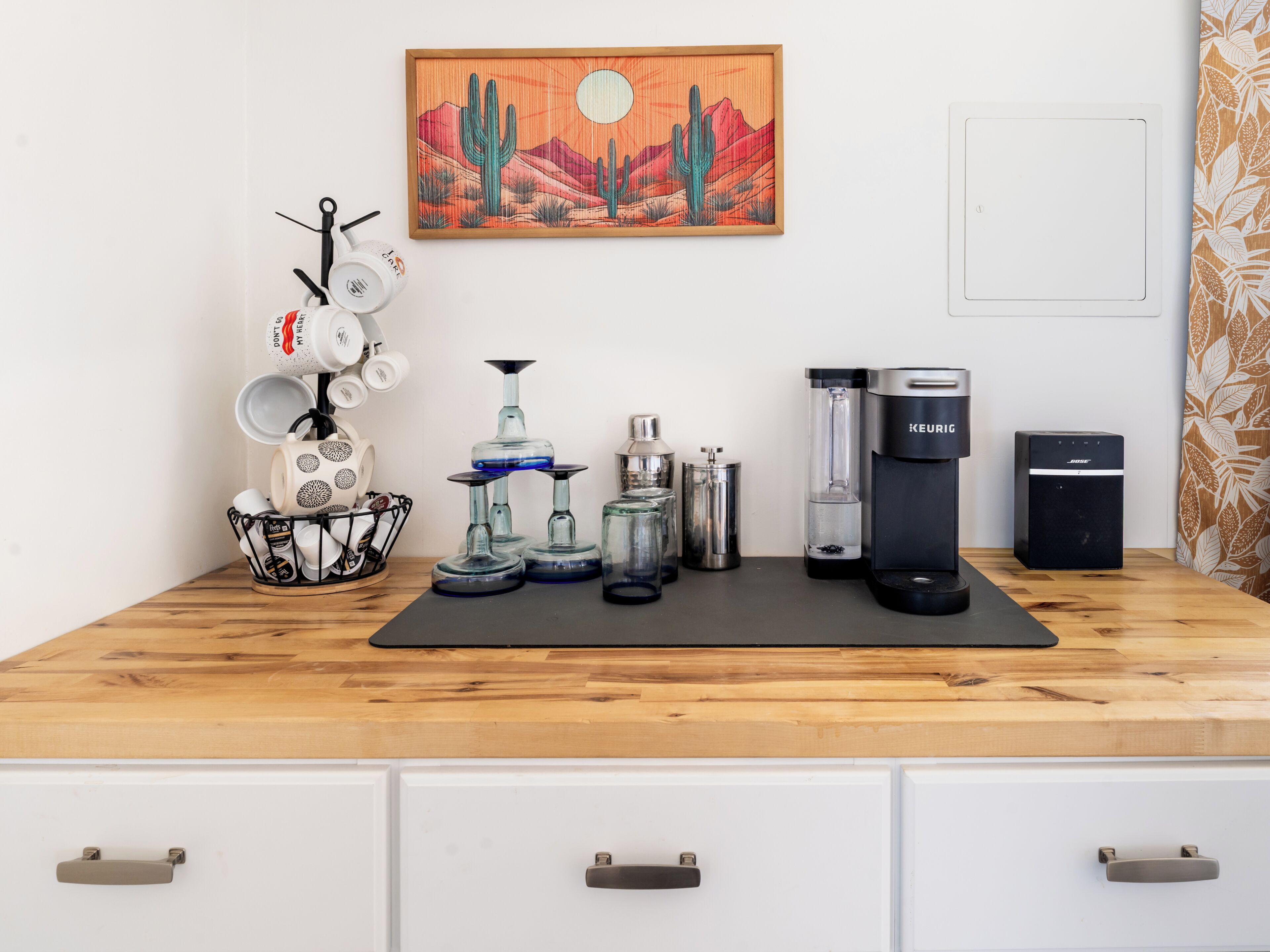A kitchen counter with a coffee maker, a Keurig, a mug rack, and a cactus painting.