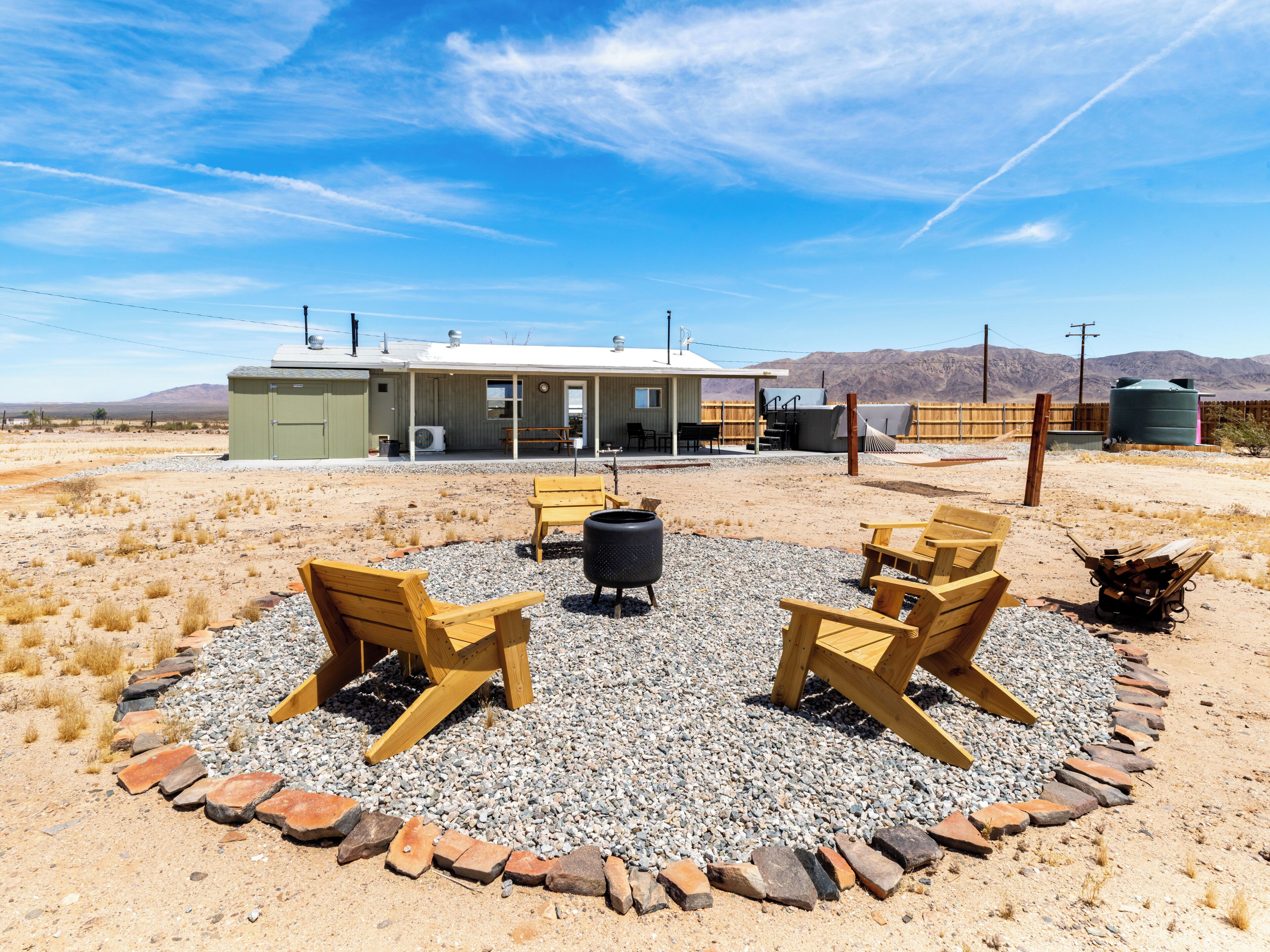 A rustic outdoor seating area with wooden chairs and a fire pit, surrounded by a gravel circle and a brick border, with a building in the background.