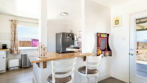 Cocina compacta con encimera de madera, dos sillas blancas, un refrigerador, un bote de basura y una puerta que da al exterior.