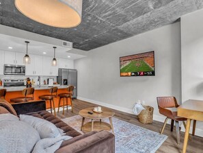 A modern living room with a sofa, coffee table, and a TV mounted on the wall displaying a sports game. - The Summit Nest | Cozy 1BR in the Heart of Knox (Knoxville)