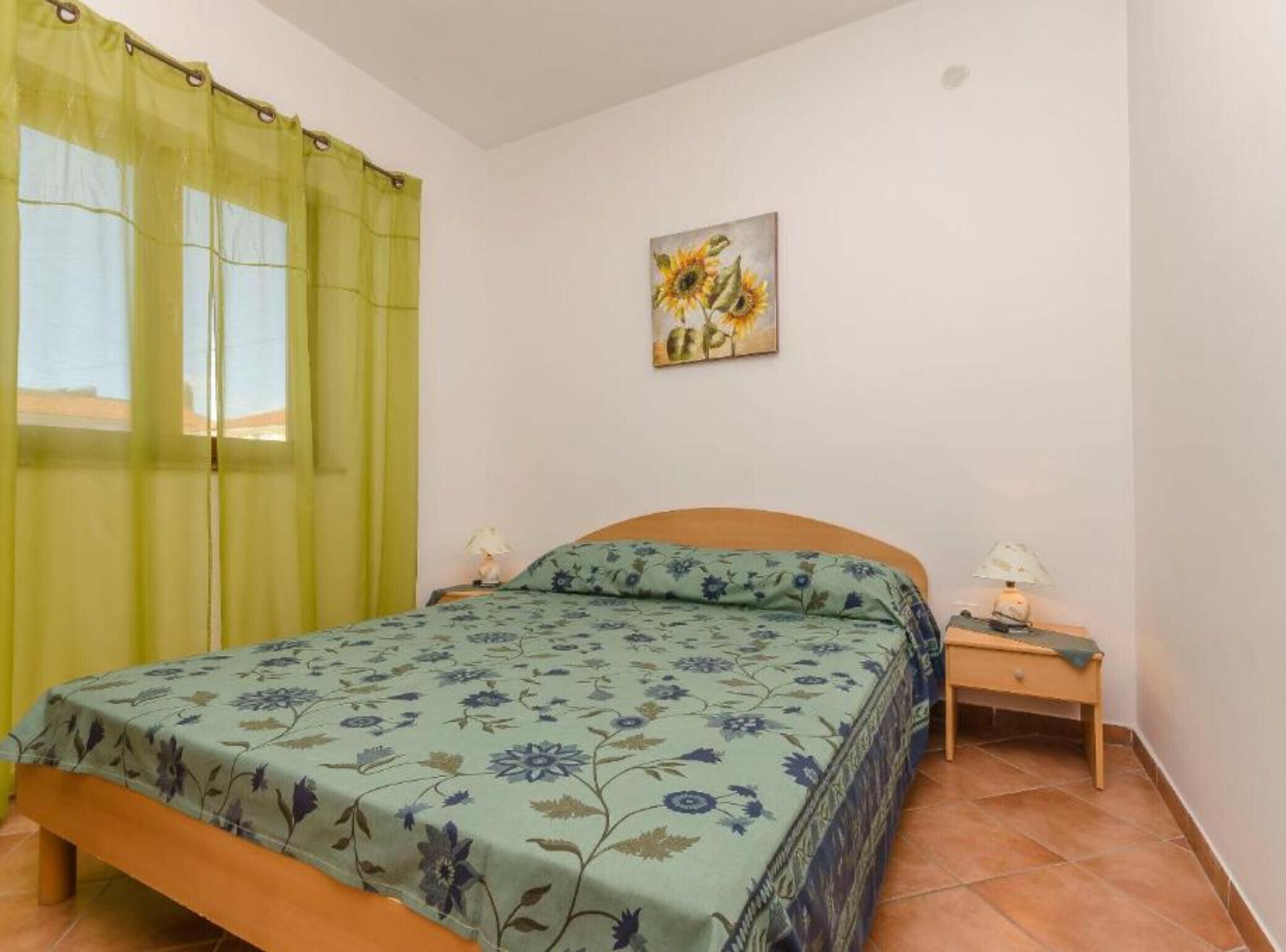A bedroom with a bed, a nightstand, a lamp, a painting, and a window with curtains.