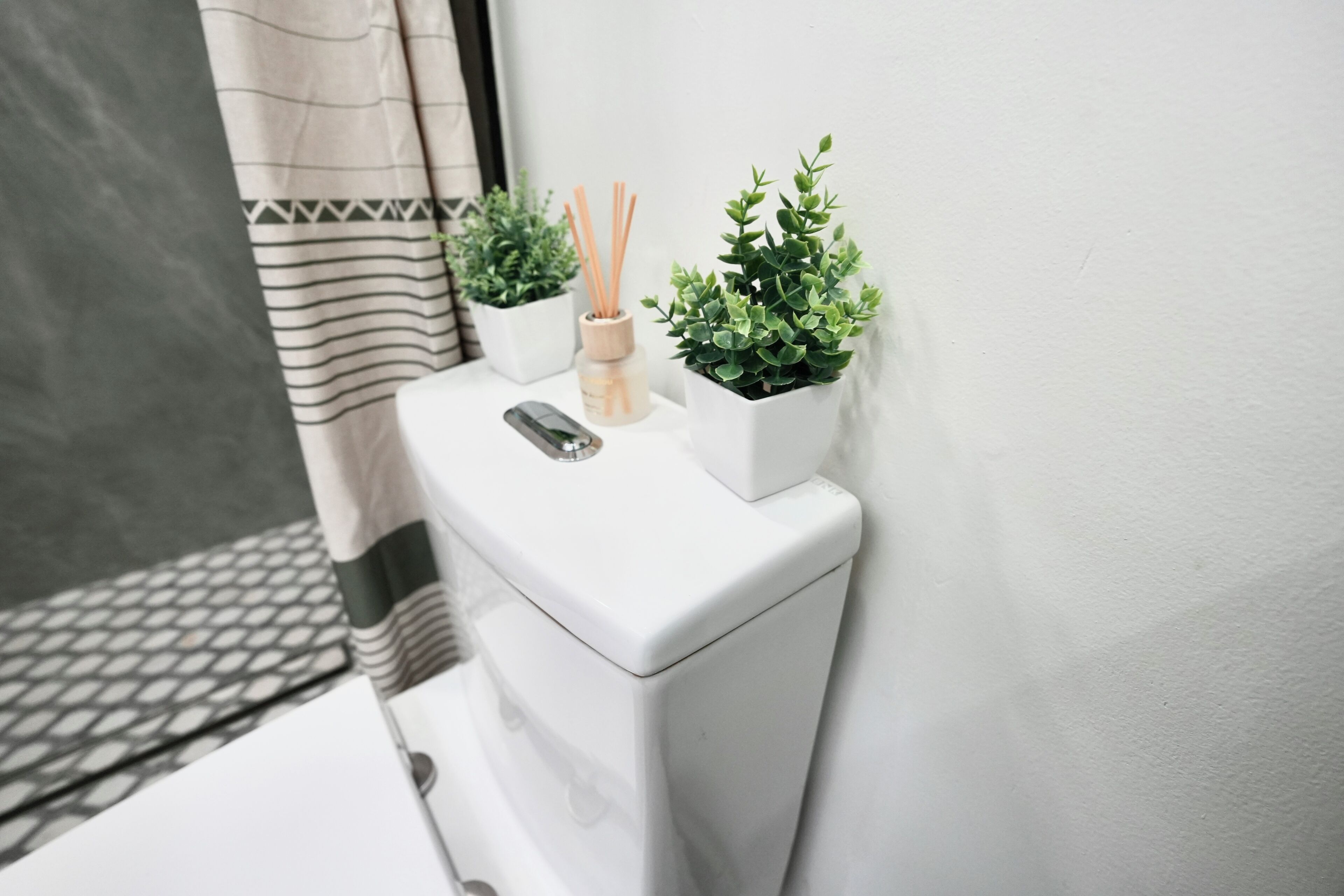 A white toilet with a remote control, two potted plants, and a small bottle of reed diffuser.