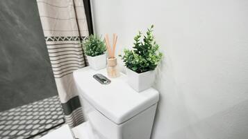 A white toilet with a remote control, two potted plants, and a small bottle of reed diffuser.