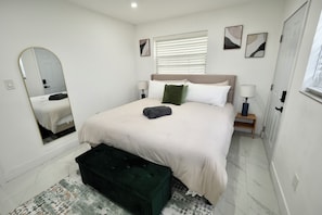 A bedroom with a bed, a mirror, a nightstand, a lamp, a window with blinds, and a door. - 6370 Atlanta House (Hollywood)