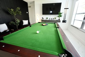 Une table de billard recouverte d’un drap de feutre vert, une boule blanche et un jeu de billes colorées. Un canapé noir avec des coussins blancs et une plante en pot se trouvent à gauche. Un téléviseur à écran plat est fixé au mur, au-dessus de la table de billard.