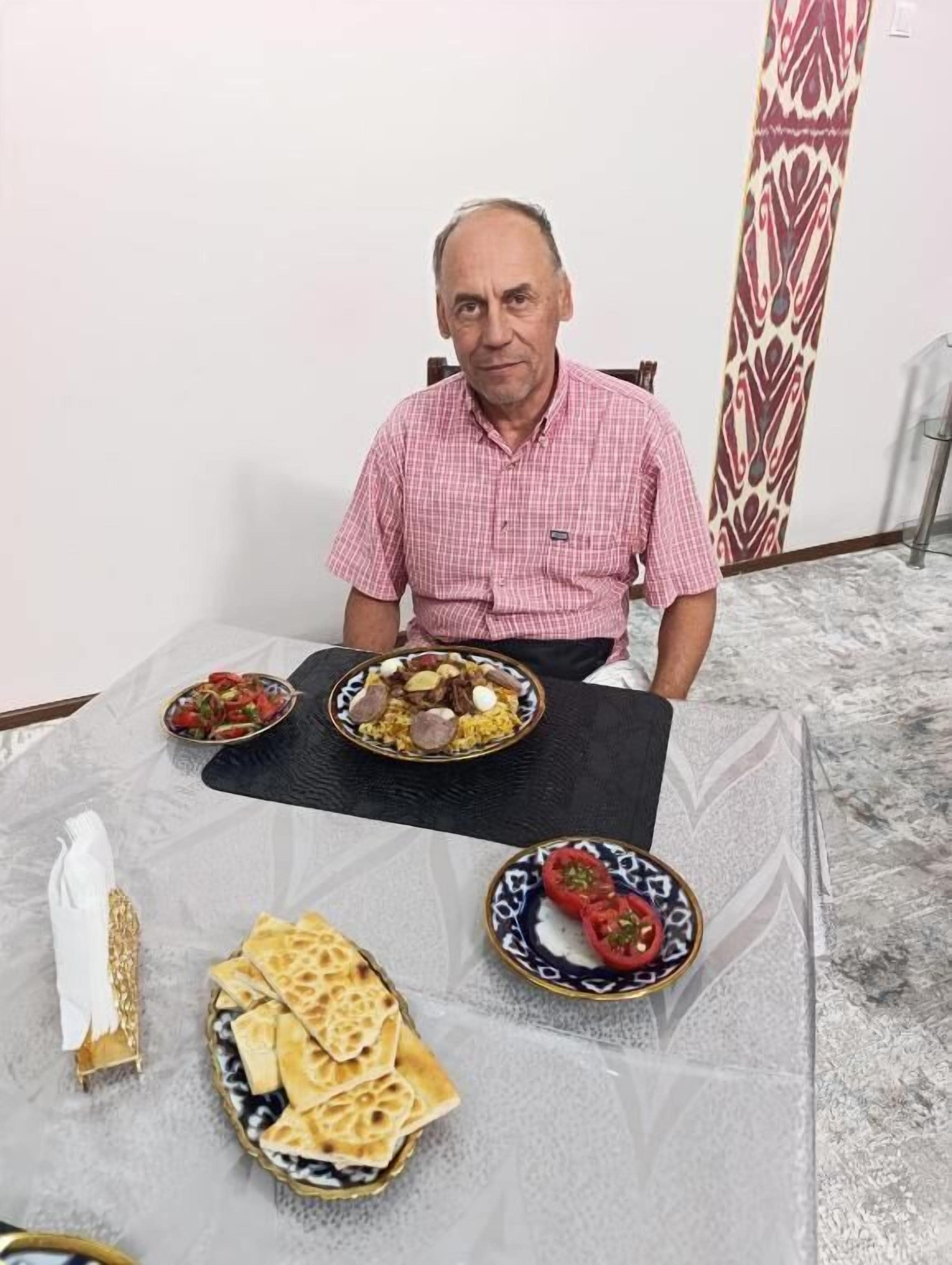 A man seated at a table with various dishes, a patterned wall, and a patterned curtain in the background.