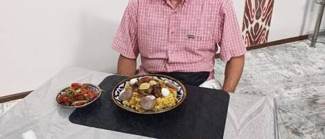A man seated at a table with various dishes, a patterned wall, and a patterned curtain in the background.