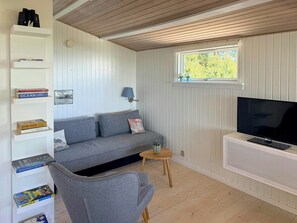 A cozy living room with a sofa, a chair, a TV, a bookshelf, and a window with a view of greenery.