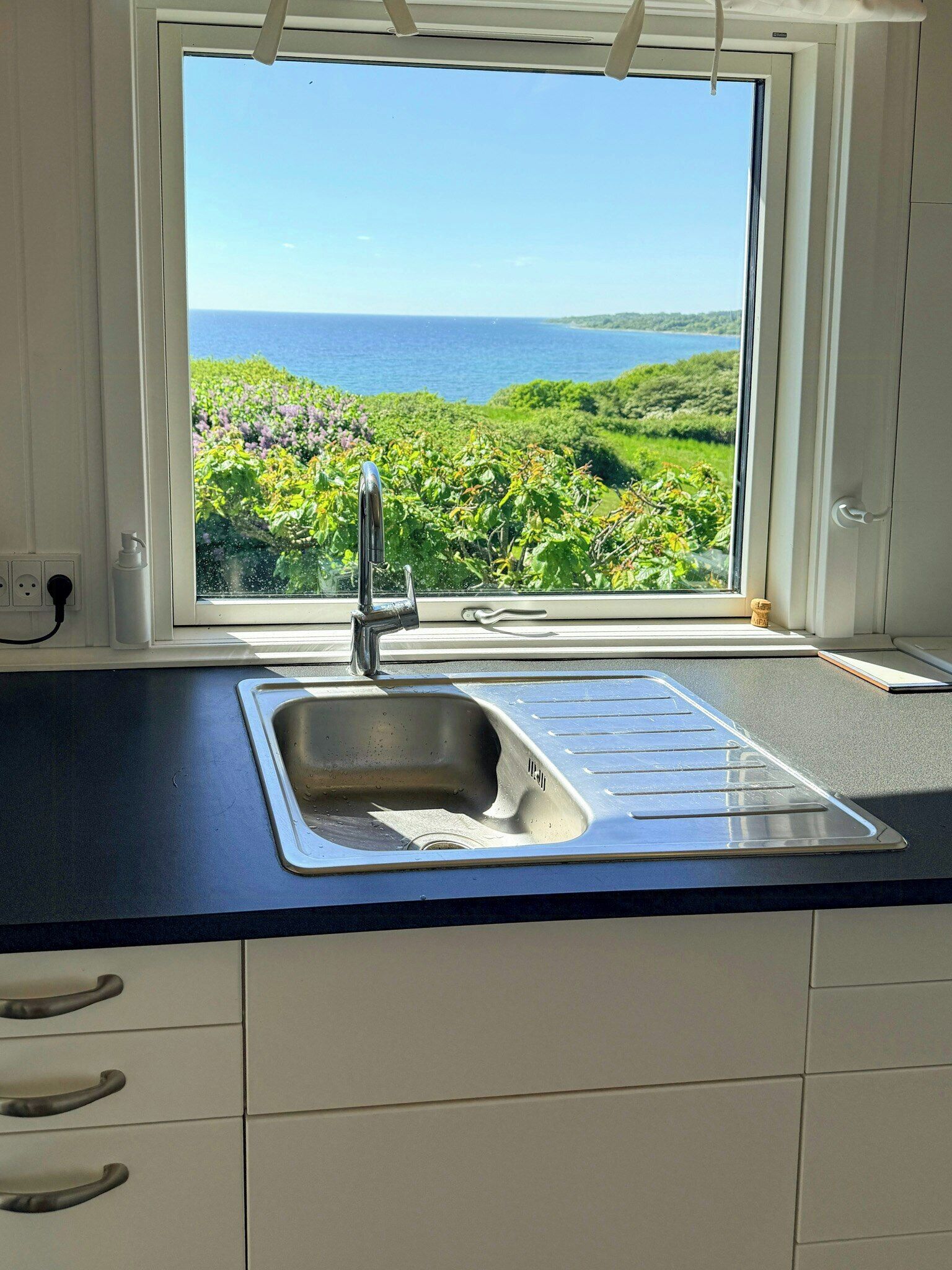 Una cocina con vista al paisaje costero, un lavamanos de acero inoxidable y una encimera blanca.