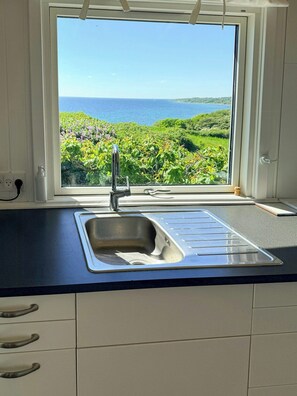 A kitchen with a window view of a coastal landscape, a stainless steel sink, and a white countertop.