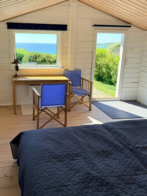 A bedroom with a bed, a desk, two chairs, a window with a view of the sea, and a door leading outside.