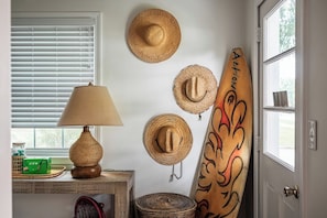 A room with a lamp, a surfboard, and several woven hats on the wall. - Cheeky Tiki | Walk to Sound Beach, Fire Pit, Hammock & Fenced Yard (Morehead City)