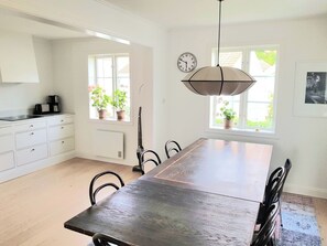 Una cocina con mesa de madera, armarios blancos y plantas en macetas junto a las ventanas.