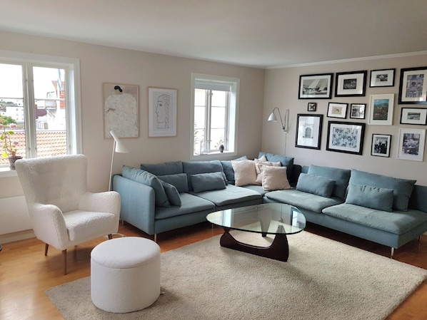 A living room with a sectional sofa, a round glass coffee table, a white armchair, and a large window with a view of buildings.
