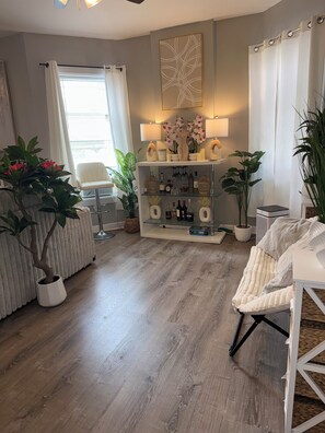 A modern living room with a wooden floor, a white armchair, a glass coffee table, and a decorative shelf with plants and bottles. -  The Langley Luxe Modern style unit (Chicago)