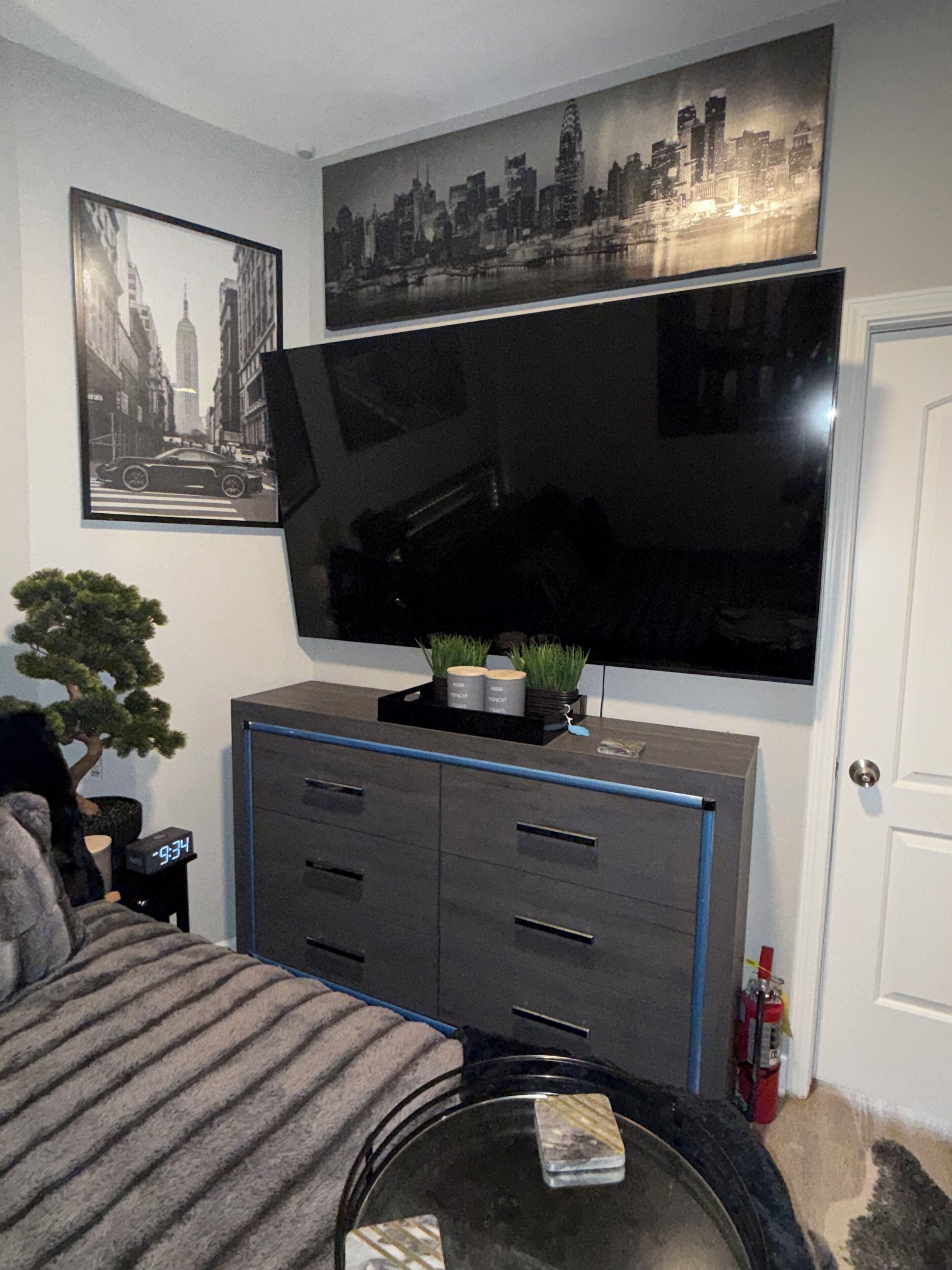 Una sala moderna con un televisor de pantalla plana montado en la pared, un mueble oscuro con cajones, una pequeña mesa con cubierta de vidrio y una planta en maceta.