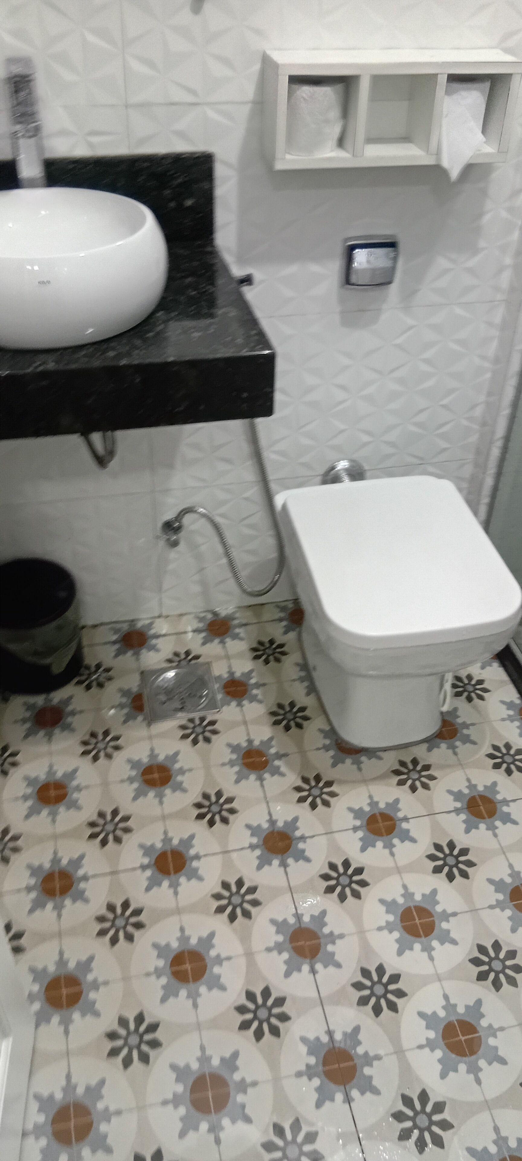Une salle de bain avec des toilettes blanches, un lavabo doté d’un plan de travail noir et des carreaux de sol à motifs.