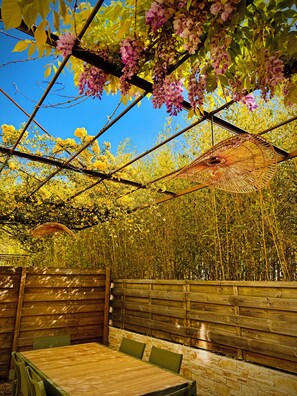 Ein Sitzbereich im Freien mit einem Holztisch und -stühlen, umgeben von einem Bambuszaun und einem mit blühenden Weinranken bewachsenen Pergola.