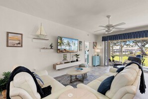 A living room with a sofa, a coffee table, a TV, a sailboat decoration, and a view through a large window.