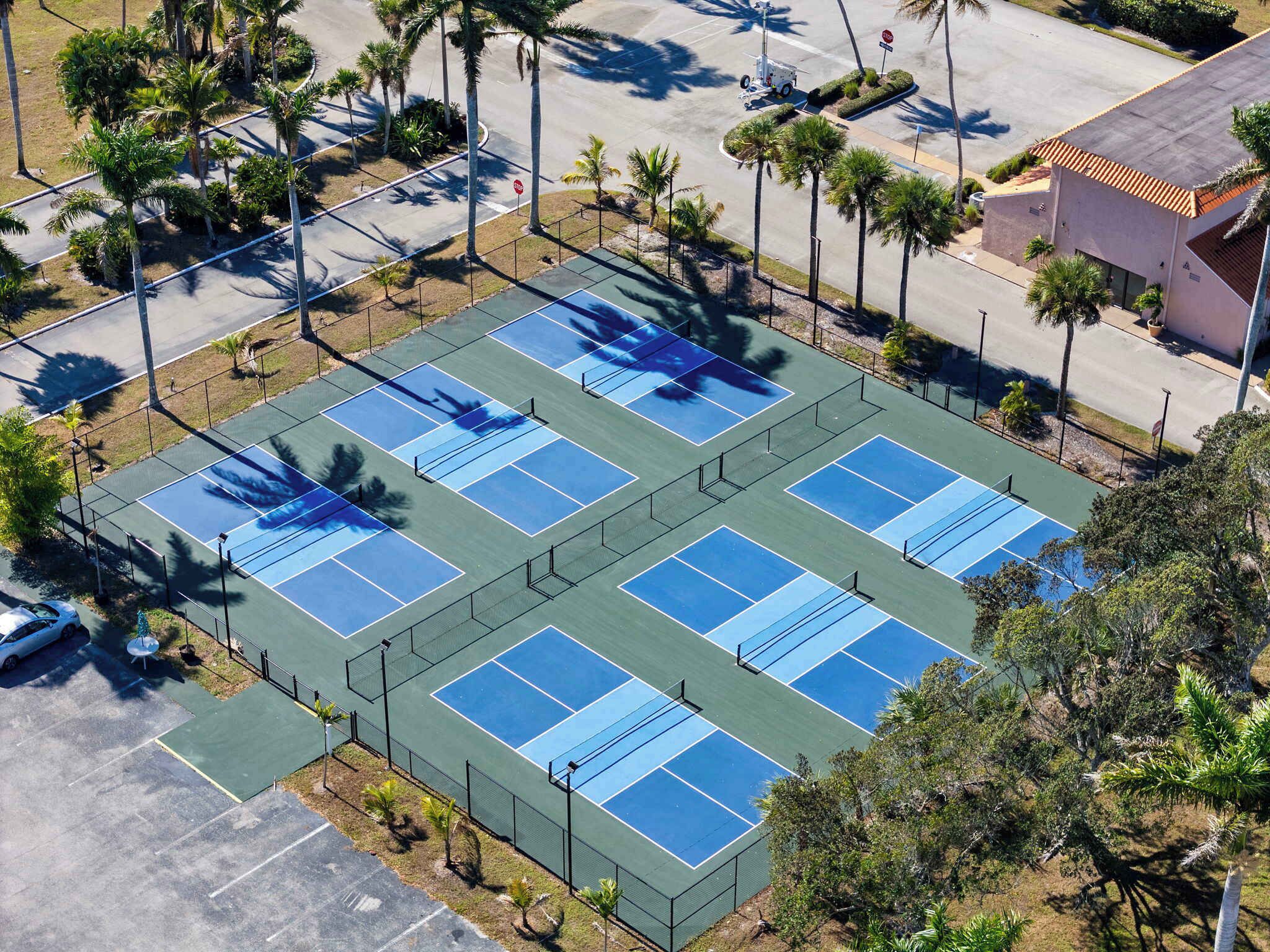 An aerial view of a tennis court complex with multiple courts, surrounded by palm trees and a parking area.