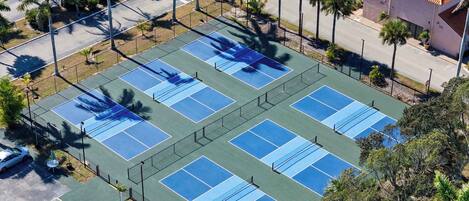 An aerial view of a tennis court complex with multiple courts, surrounded by palm trees and a parking area.