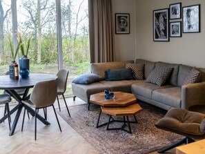 Ein modernes Wohnzimmer mit einer Couch, einem Couchtisch und Sesseln. An den Wänden hängen Bilder und durch das Fenster ist eine Baumlandschaft zu sehen.