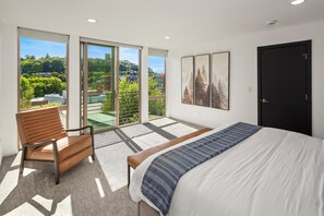 Un dormitorio con cama, una silla, un balcón con vistas a zonas verdes y edificios, y una puerta.