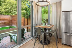 Una cocina con una mesa de madera, sillas metálicas, un refrigerador de acero inoxidable y vistas a un jardín a través de amplios ventanales.