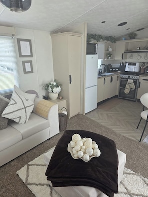 A compact living space with a sofa, a coffee table with decorative items, a kitchen area with appliances, and a dining area. - Waterside Retreat T21 (Ingoldmells)