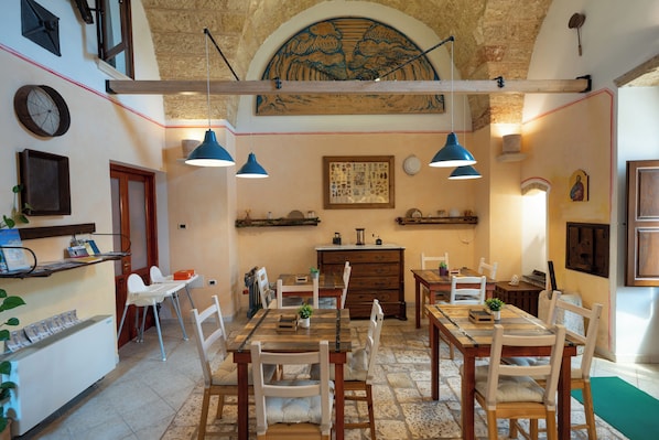 A dining area with wooden tables and chairs, a chandelier, and a stone-tiled floor.