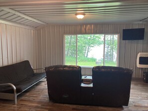 Et værelse med trægulv, en sofa, to lænestole, et bord med fjernbetjening, et fjernsyn og et vindue med udsigt over grønne områder.