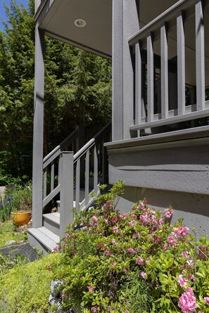 Eine graue Treppe führt zu einer Veranda mit Geländer, umgeben von Grünpflanzen und rosa Blüten.
