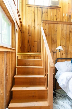 Eine Holztreppe führt zu einem Bett in einem Raum mit Holzwänden und einem Fenster.