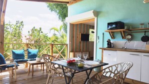 A wooden deck with a table and chairs, a kitchen area with cabinets and a microwave, and a view of greenery outside. - Caribbean Beach Studios. Ti Colibri Studio near the beaches in Ferry-Deshaies (Deshaies)