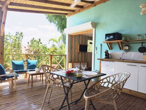 Une terrasse en bois avec une table et des chaises, un coin cuisine équipé de placards et d’un four à micro-ondes, et une vue sur la verdure environnante.