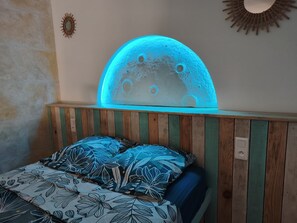 Ein Schlafzimmer mit einem Holz-Kopfteil, einem Bett mit blumigem Bettbezug und einer beleuchteten Monddeko an der Wand.