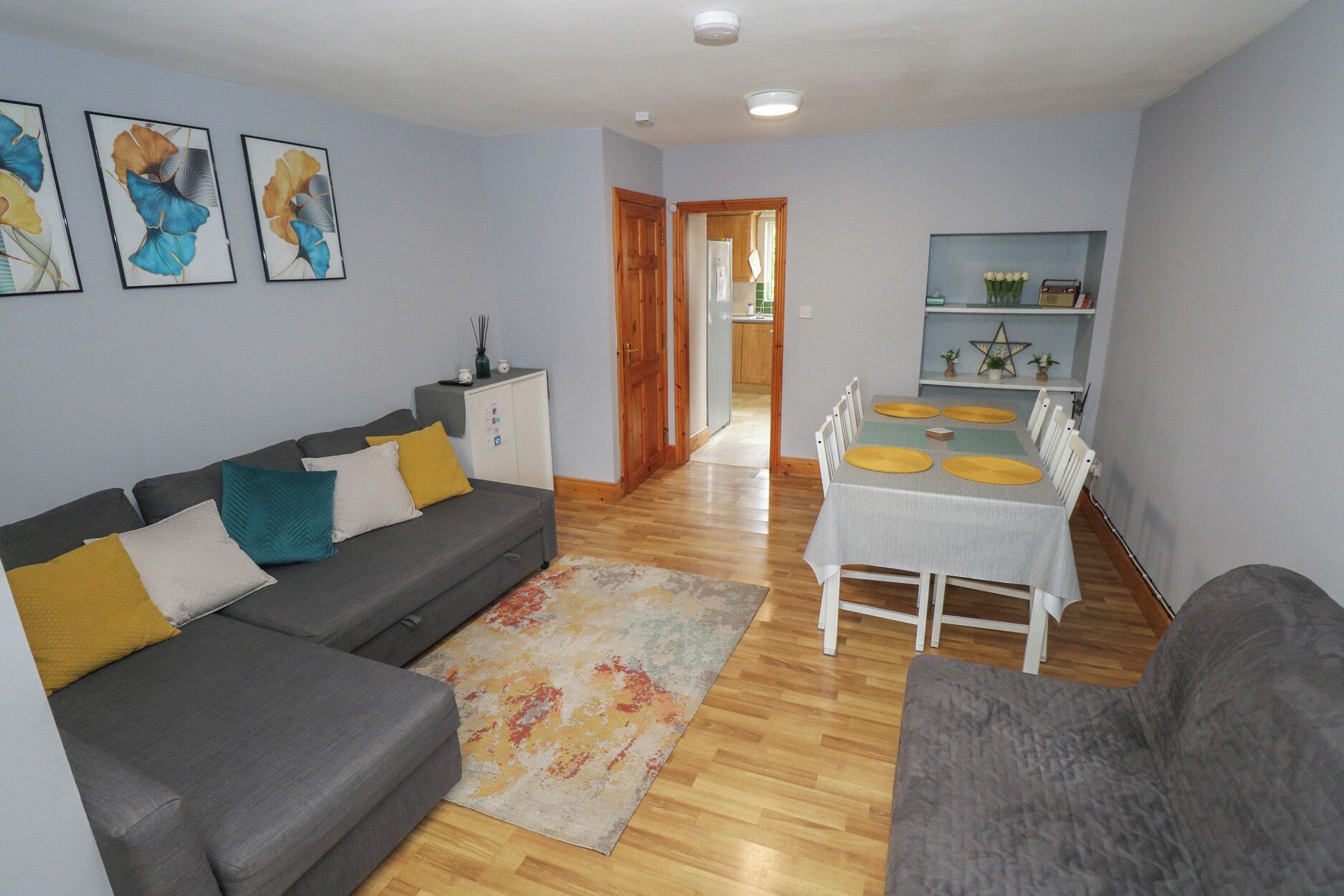 A living room with a grey sofa, a dining table with yellow placemats, and a kitchen in the background.