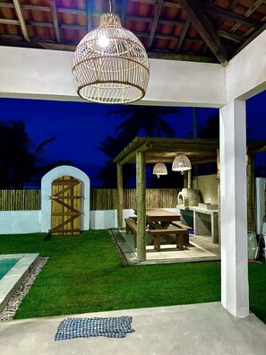 Ein überdachter Außenbereich mit einer Holzpergola, Picknicktischen, einem Pool und einer hängenden Leuchte.