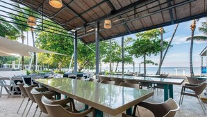 An outdoor dining area with tables and chairs, a thatched roof, and a view of the ocean. - Emotions by Hodelpa - Juan Dolio (Juan Dolio)