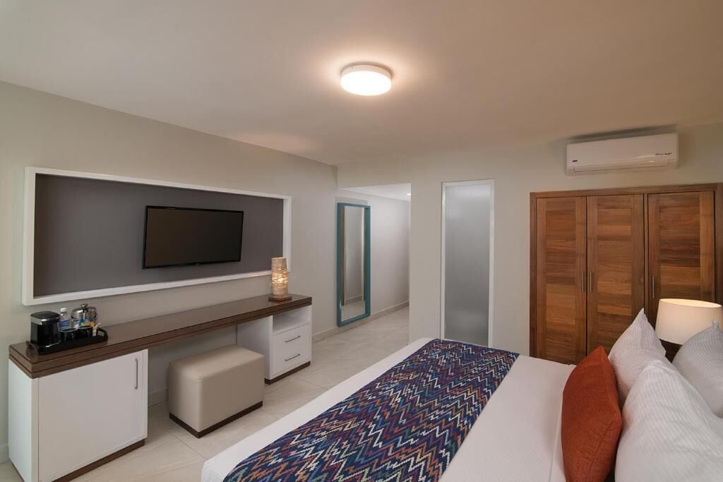 A hotel room with a bed, a TV mounted on the wall, a desk with a chair, a wardrobe, and an air conditioning unit.