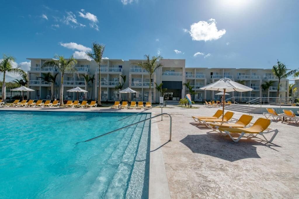 Un área de piscina con tumbonas y sombrillas, un edificio con balcones y palmeras.