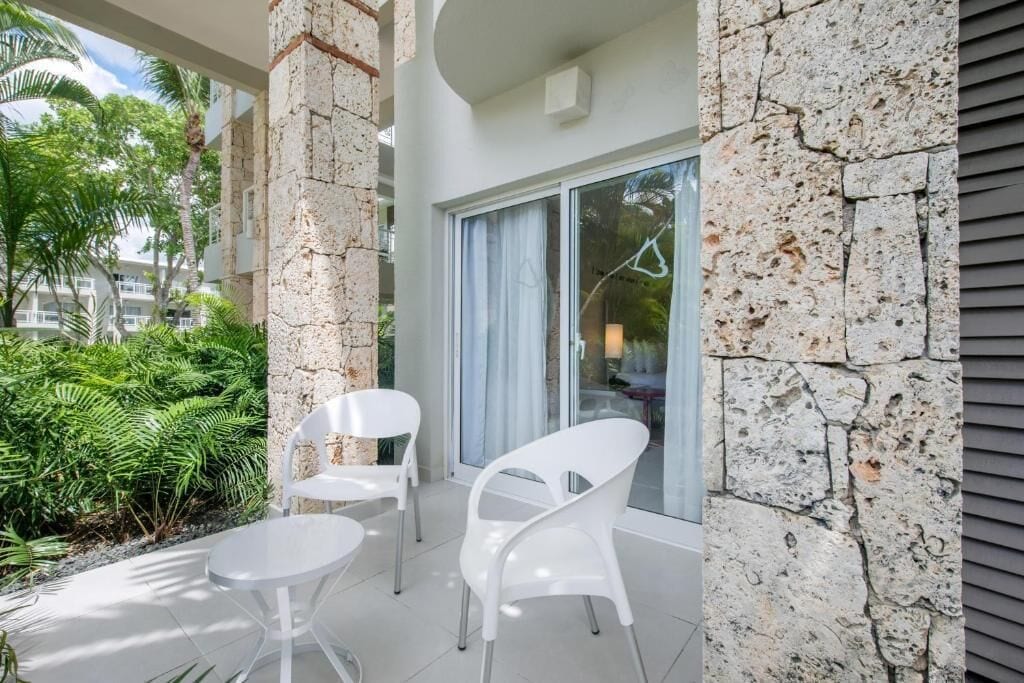 A balcony with white chairs and a table, stone walls, and a view of greenery.