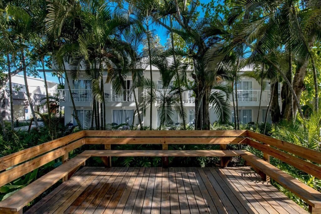Una terraza de madera con bancos, rodeada de exuberante vegetación y palmeras, con vistas a un edificio blanco con balcones.