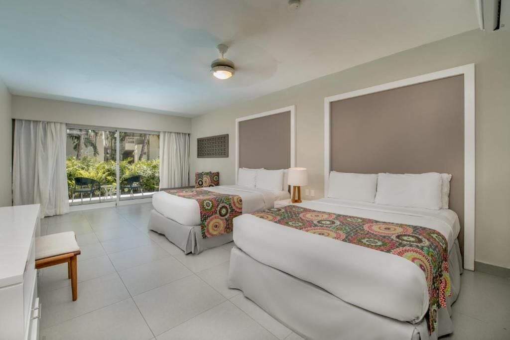 A hotel room with two beds, a desk, and a view of greenery outside.