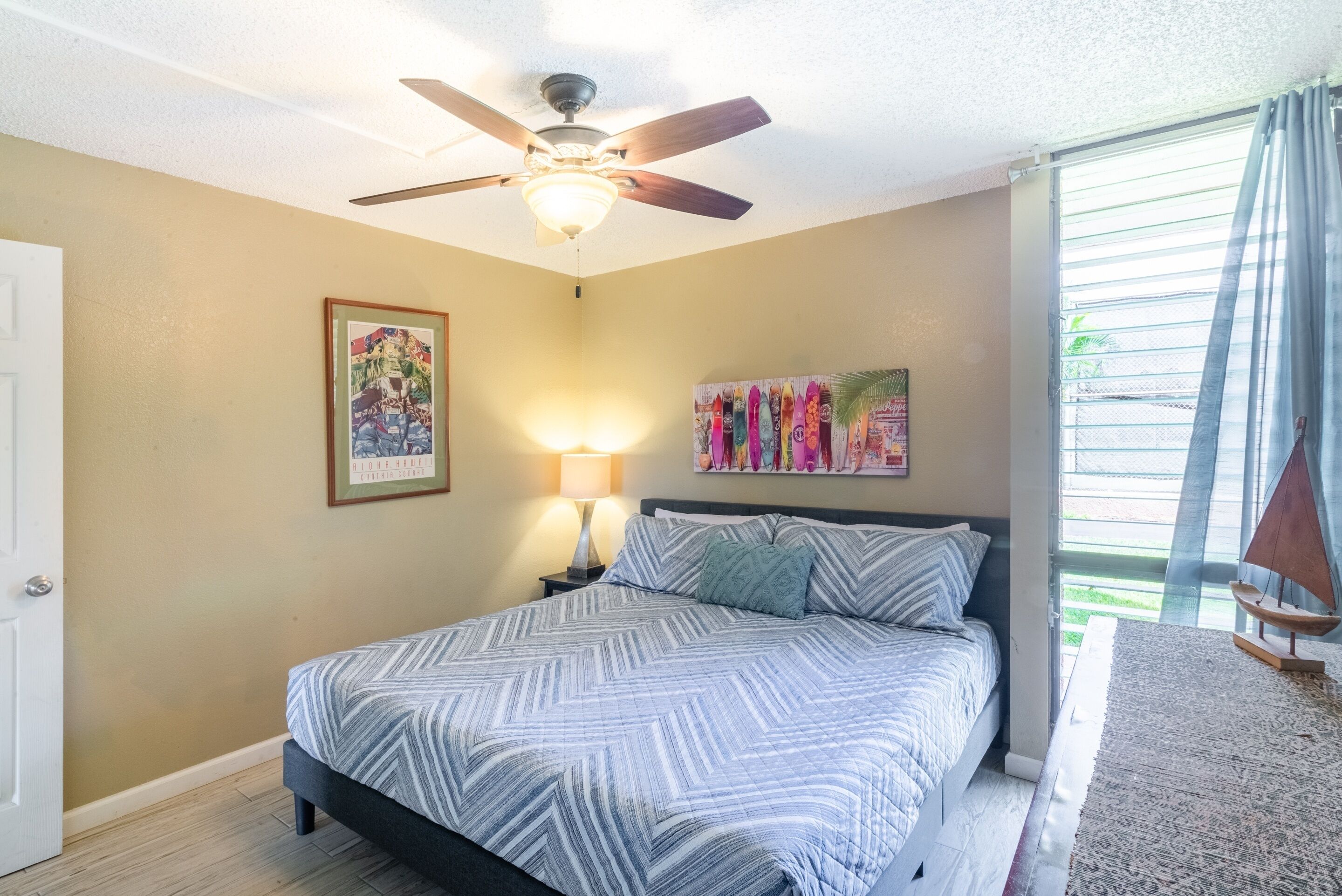 A bedroom with a bed, a ceiling fan, a nightstand with a lamp, and artwork on the walls.