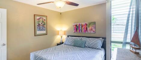 A bedroom with a bed, a ceiling fan, a nightstand with a lamp, and artwork on the walls.