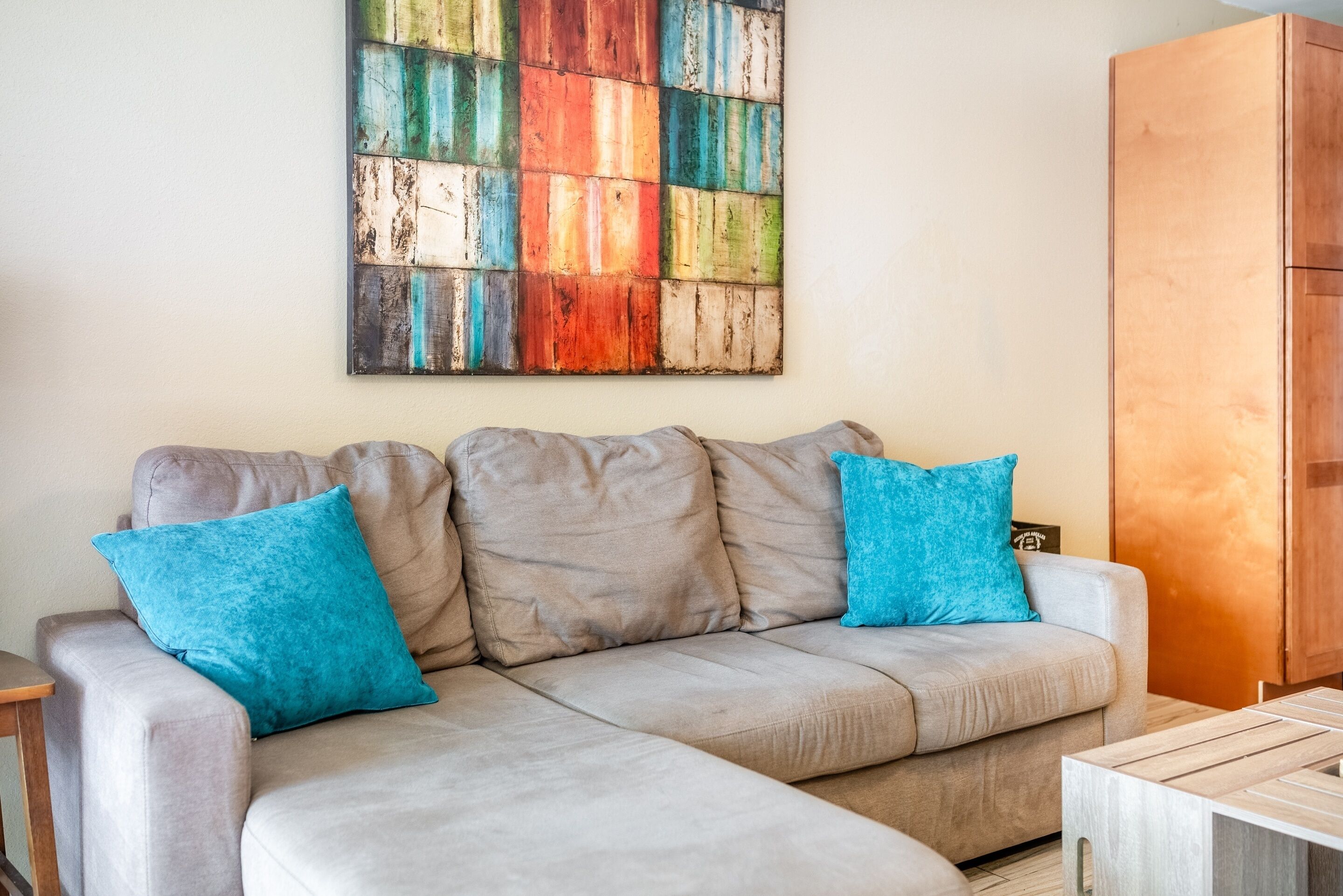 A beige sectional sofa with two blue cushions, a wooden coffee table, and a colorful abstract painting on the wall.