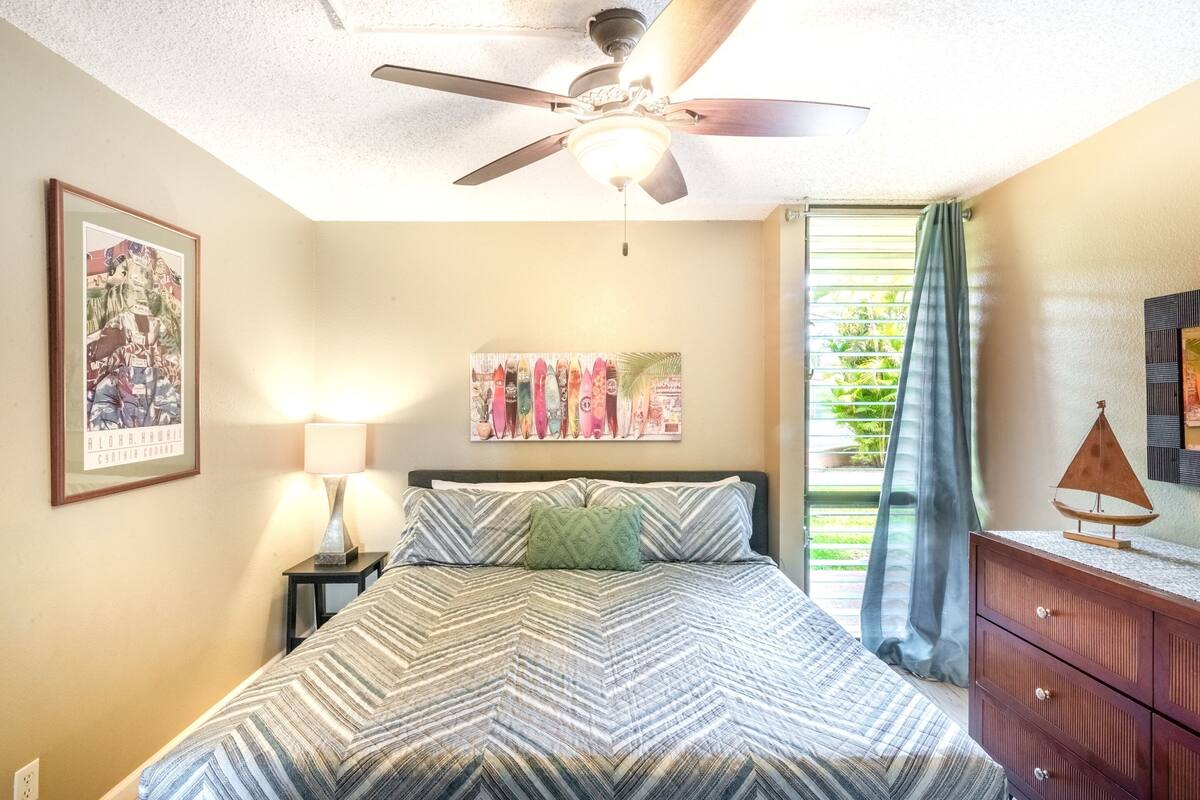 A bedroom with a bed, a ceiling fan, a nightstand with a lamp, a framed picture, a colorful artwork, and a view of greenery through a window.
